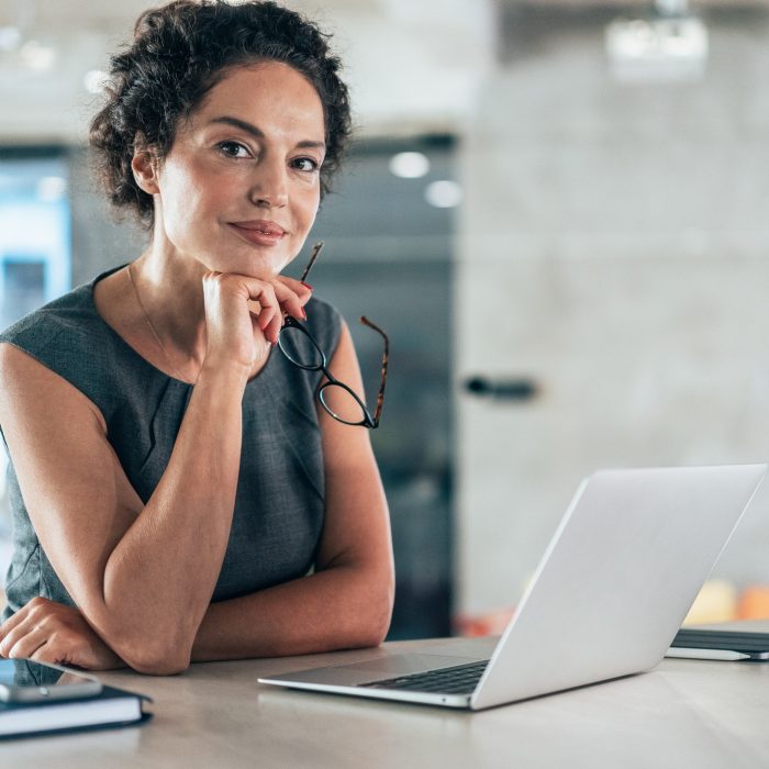 Confident mid age businesswoman working on laptop at her workplace at modern office. Portrait of businesswoman in front of her laptop, looking at camera and smiling