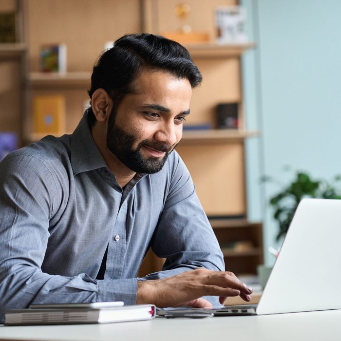Smiling indian business man working on laptop at home office. Young indian student or remote teacher using computer remote studying, virtual training, watching online education webinar at home office.